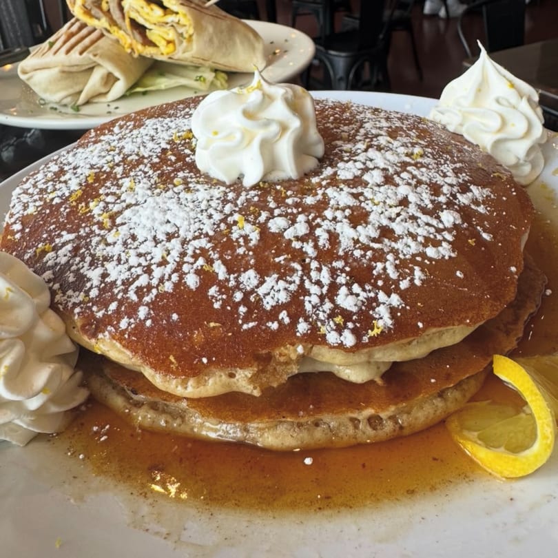 Fluffy pancakes with powdered sugar, whipped cream, lemon zest, and syrup, with a breakfast burrito in the background.