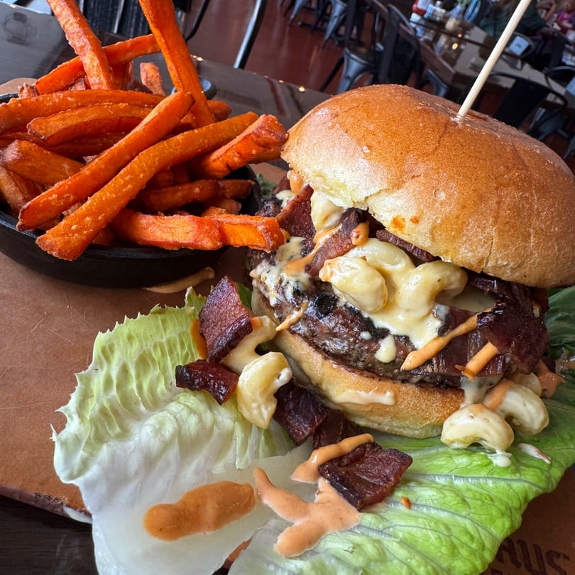 A burger topped with mac and cheese, bacon, and sauce, served with sweet potato fries.