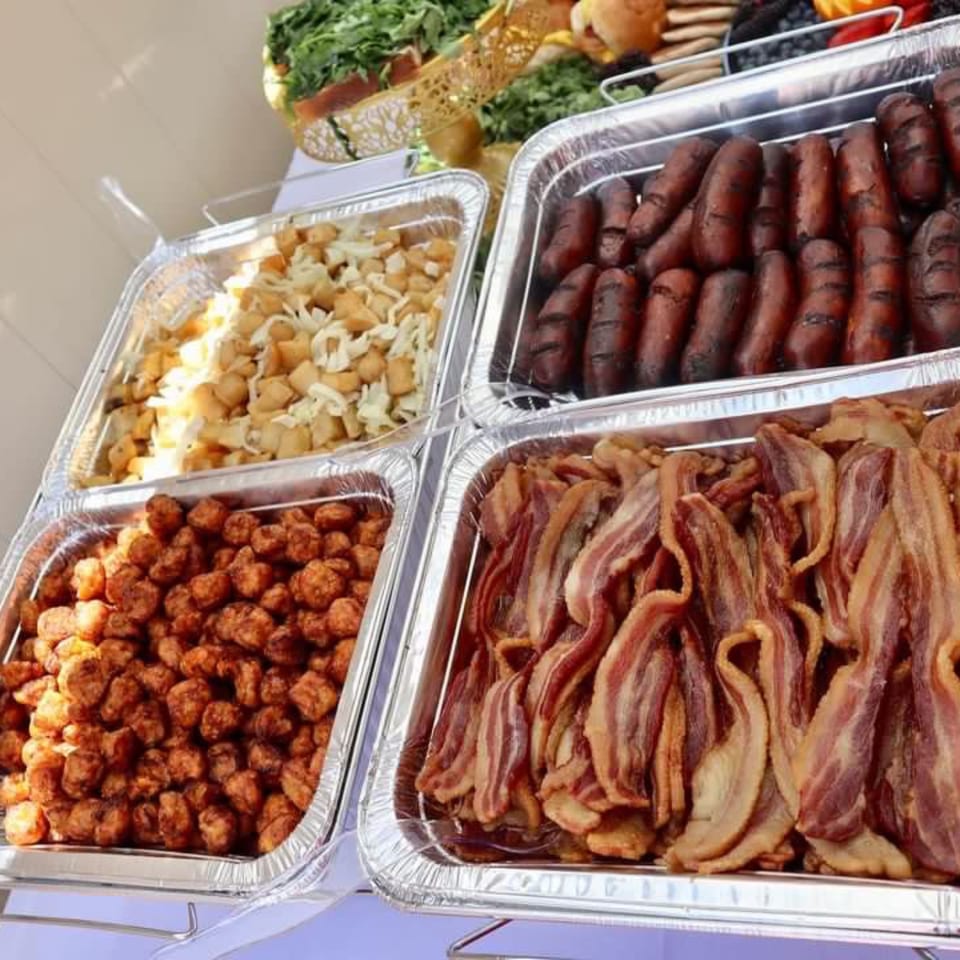 Catering trays filled with crispy bacon, sausages, breakfast potatoes, and tater tots.