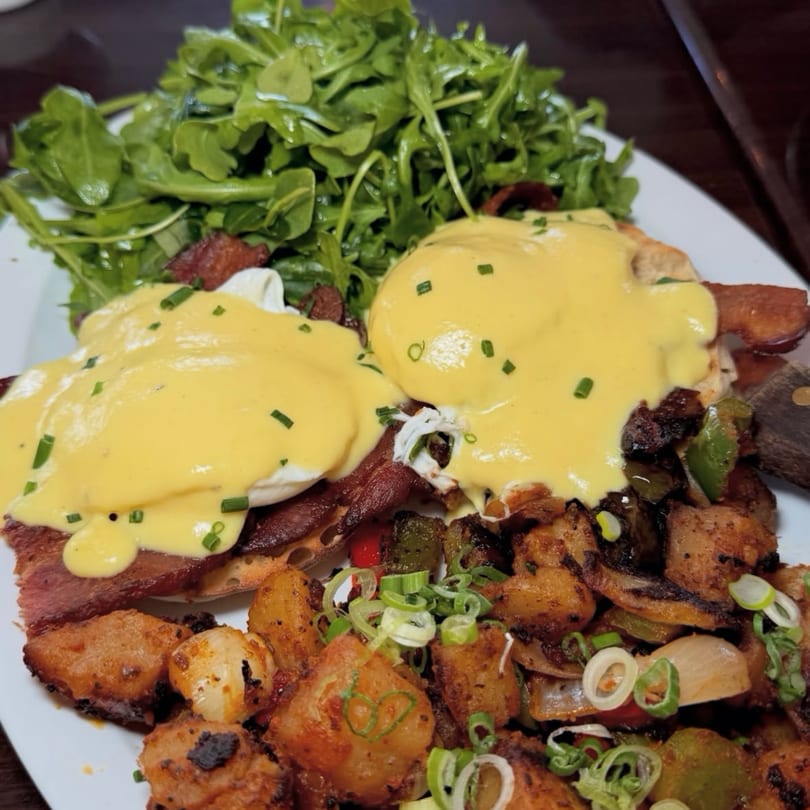 Eggs Benedict with bacon, hollandaise sauce, a side of roasted potatoes, and a fresh arugula salad.