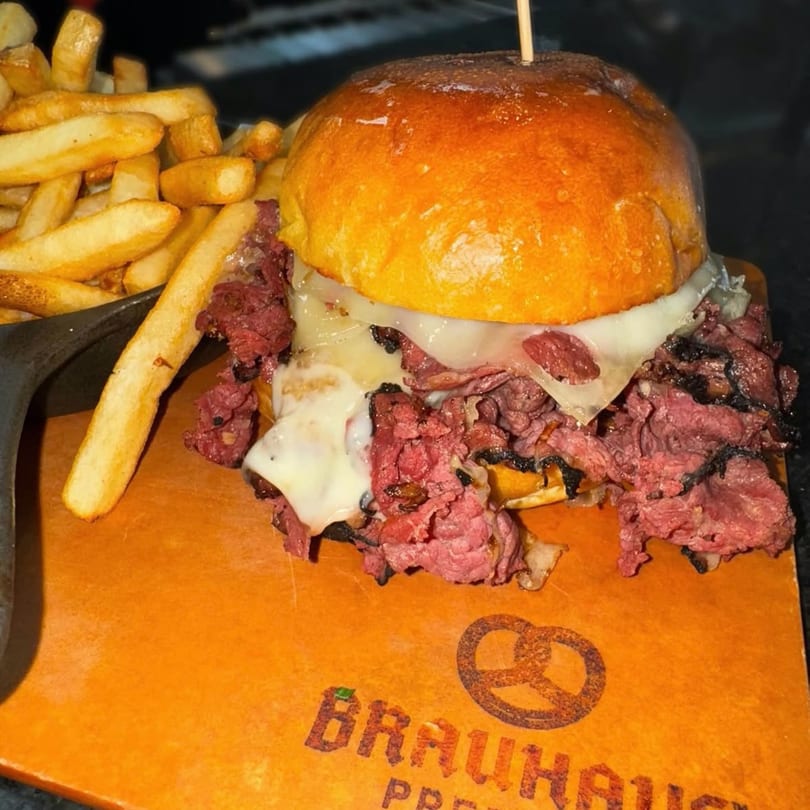 A pastrami sandwich with melted cheese and fries on a wooden board with a "Brauhaus" logo.