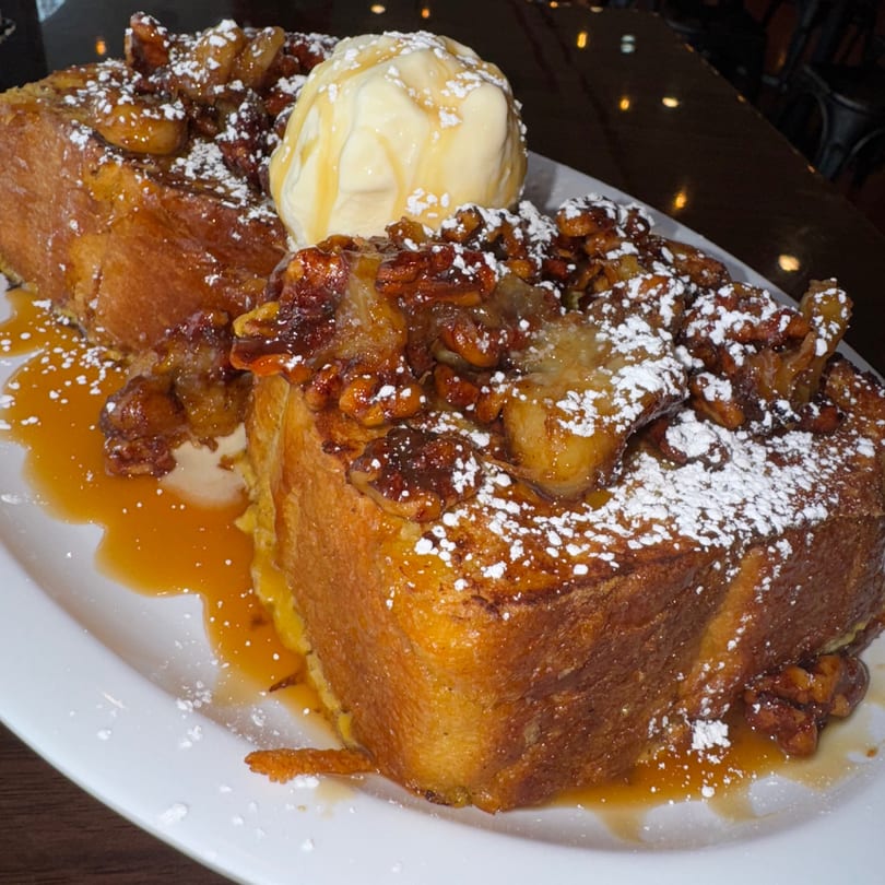Thick French toast topped with caramelized bananas, pecans, powdered sugar, syrup, and a scoop of ice cream.